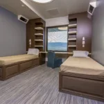 A modern room with two single beds, a central window featuring a scenic view, a blue chair, and built-in wooden shelving on either side. The walls are painted in a soothing gray with matching flooring. The room is well-lit and organized.