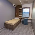 A minimalist room with a single bed, beige bedding, and a small pillow. The room has a chair, a built-in desk under a window with blinds, shelves, and neutral-colored walls and flooring. A small wall-mounted TV is visible in the corner.