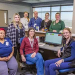 A group of eight people, including healthcare workers and staff, are gathered in a medical office. Some are sitting, and others are standing behind a desk with computer monitors and medical equipment. They are smiling and appear to be part of a team.