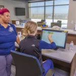 A healthcare professional in blue scrubs consults with a colleague seated at a computer in a modern office. The seated person points at the screen, and a water bottle is on the desk. The room has large windows and a relaxed atmosphere.
